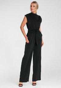 Czarny kombinezon z krótkimi rękawami, elastycznym pasem i zawiązywanym paskiem. Miękka tkanina, szerokie nogawki i minimalistyczny design. Czarne sandały na obcasie z odkrytymi palcami.