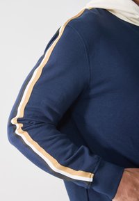 Felpa blu navy con cappuccio foderato in beige, caratterizzata da audaci righe dorate, bianche e nere lungo le maniche. Tessuto morbido in cotone.