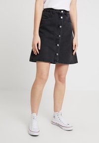 Černá džínová sukně s rozšířeným tvarem, zapínání na přední knoflíky a designem s pěti kapsami, kombinovaná s bílými vysokými teniskami.