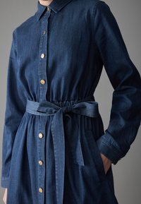 Jeanskjolklänning i mörkblått, med krage, guldknappar, elastisk midja och knytbälte. Två sidofickor synliga.