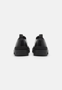 Chaussures en cuir noir à forme arrondie, avec des semelles épaisses en caoutchouc et des tirettes en tissu à l'arrière, présentant une texture lisse et un design minimal.