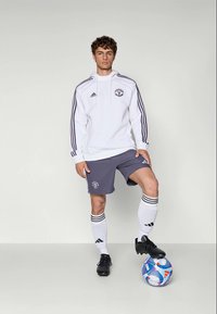 Haut à capuche blanc avec des rayures sombres, short gris, chaussures de foot noires et chaussettes blanches avec des rayures noires. Un ballon de football aux pieds, présentant un design coloré.