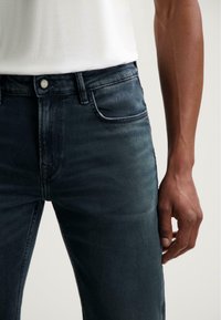 Tmavě modré džínové kalhoty se slim fit střihem, s klasickým designem pěti kapes a stříbrným knoflíkem v pase. Hladká textilie.