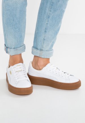 Hvide lædersneakers med tykke brune såler båret med opkrogede lyseblå jeans på en ensfarvet hvid baggrund.