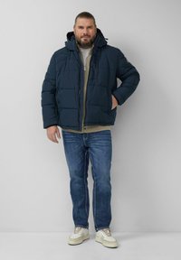 Granatowa puchowa kurtka z kapturem, z pikowaną fakturą i zamkiem z przodu, zestawiona z beżową bluzką, niebieskimi dżinsami i białymi trampkami.