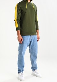 Uomo che indossa un hoodie verde oliva con strisce gialle sulle maniche, jeans azzurri chiari e sneakers bianche, in piedi contro uno sfondo bianco.