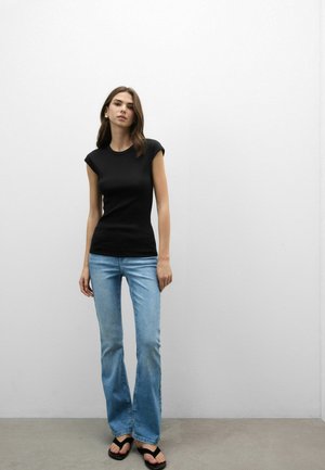 Kvinde, der står op ad en hvid væg iført sort top med korte ærmer, lyseblå vide jeans og sorte flip-flop sandaler.