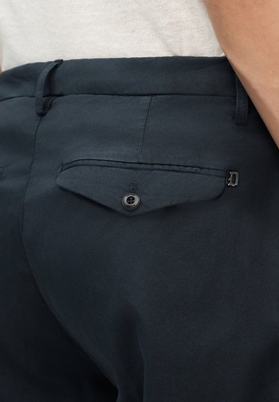 Gros plan sur la poche arrière d'un pantalon bleu foncé avec un rabat boutonné et une petite étiquette métallique en forme de D sur le côté droit.