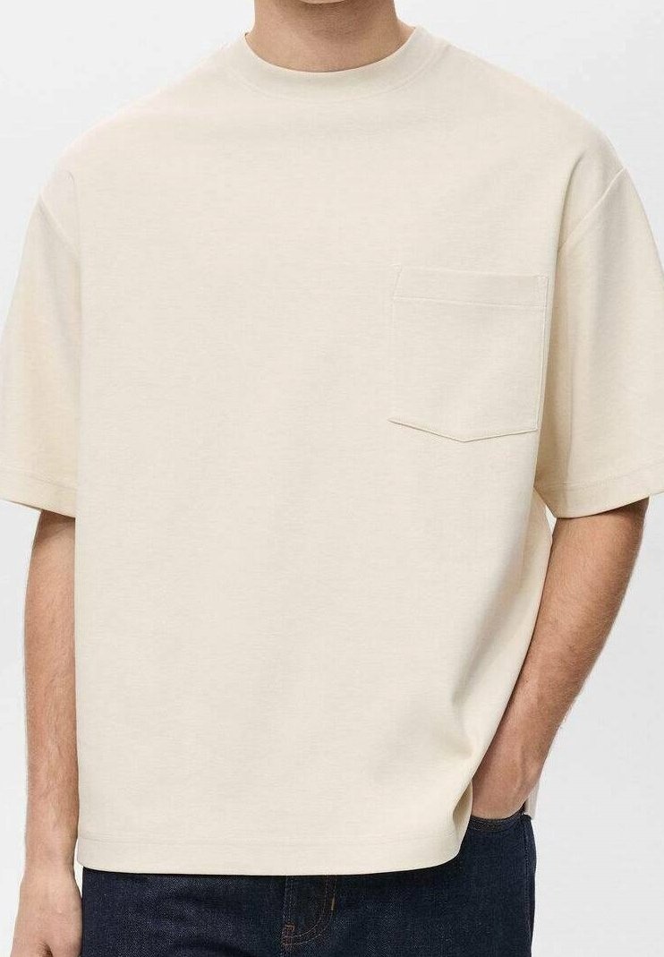 T-shirt beige à manches courtes avec une poche poitrine, porté par une personne ayant une main dans la poche d’un jean bleu foncé.