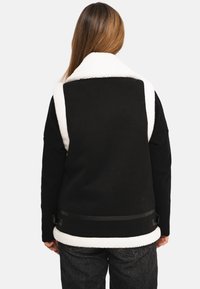 Veste sans manches noire et blanche avec un col et un dos en polaire, présentant une texture lisse et une ceinture noire ajustable à la taille.