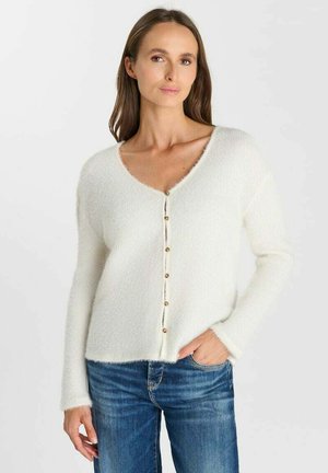 Vrouw draagt een witte, pluizige, knoopjes cardigan en blauwe jeans, staat met één hand in de zak tegen een effen lichte achtergrond.
