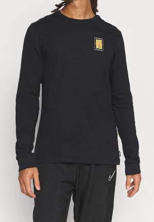 Mann trägt schwarzes Langarmshirt mit kleinem gelb-orangefarbenem Motiv auf der Brust, kombiniert mit schwarzen Nike-Hosen.