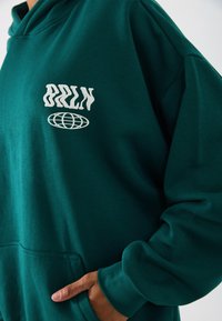 Grüne Baumwollkapuzenjacke mit Vordertasche, mit weißem "BRLN"-Text und Globe-Grafik. Weiche Textur, lockere Passform und mit Bündchen an den Ärmeln.