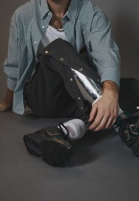 Gestreiftes grün-weißes Hemd, graue Hose mit Druckknöpfen, silbernes Prothesenbein, schwarze Sportschuhe und weiße Socken.
