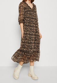 Robe maxi noire avec un design à motifs, manches bouffantes et décolleté en V. Portée avec des bottines beiges et une silhouette fluide.