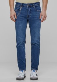 Tmavomodré denimové džínsy so slim fit strihom, dizajnom s piatimi vreckami, kovovými doplnkami a reťazovým detailom pripevneným na prednej strane.