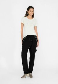 Vitt t-shirt med korta ärmar och präglad logotyp, kombinerad med svarta byxor med ränder på sidorna och metalliska klackar med ormmönster.