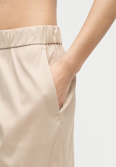 Μπεζ παντελόνι με ελαστική μέση και ένα χέρι που στηρίζεται στην δεξιά πλαϊνή τσέπη σε απλό λευκό φόντο.