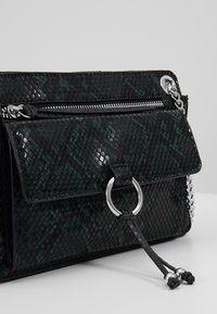 Borsa nera e verde con trama testurizzata dal motivo simile a quello di un rettile, cerniere argentate, un anello circolare decorativo e una tracolla a catena.