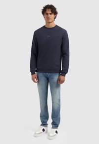 Marineblauwe trui met ronde hals, ribgebreide manchetten en onderrand; heeft kleine witte tekst "PURE PAST" op de borst. Draag het met lichtblauwe jeans en witte sneakers.