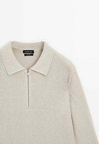 Jasnobeżowy sweter na zamek z kołnierzykiem, dzianinową fakturą i żeberkowymi detalami, z metką wskazującą na 100% bawełnę.