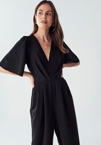 Ženska z dolgimi rjavimi lasmi, oblečena v črn kombinezon z globokim V-izrezom in širokimi rokavi, stoji s poudarjenimi boki proti beli ozadju.