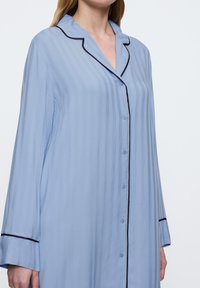 Camicione da notte blu chiaro, a maniche lunghe, con colletto, strisce verticali, piping nero lungo il colletto e la pattina dei bottoni, e chiusura a bottoni.