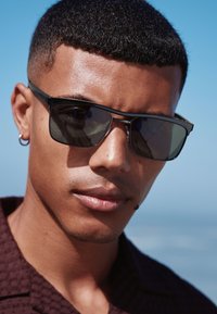 Occhiali da sole neri con montature rettangolari, lenti grigie e dettagli in metallo, indossati da una persona su uno sfondo blu.