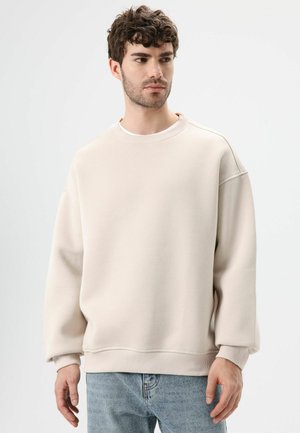 Beige sweater gemaakt van zachte stof, met een ronde hals, verlaagde schouders en geribbelde boorden. Gecombineerd met lichtblauwe jeans.