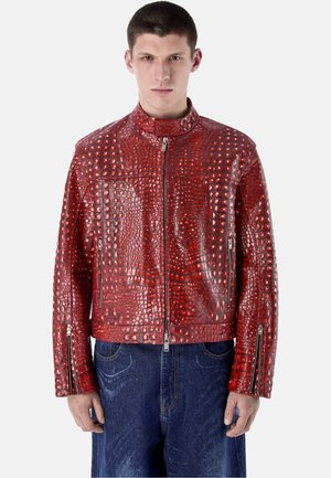 Junger Mann trägt eine glänzende rote Lederjacke mit Krokodilmuster, Reißverschlusstaschen und blaue Jeans.