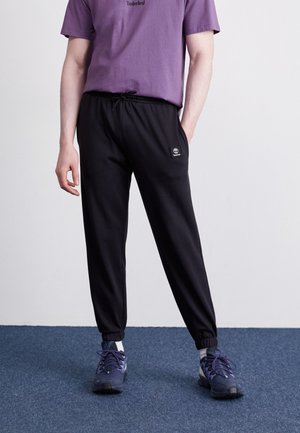 Person iført en lilla Timberland t-shirt, sorte joggers med snøre og Timberland-logo, og mørkeblå sneakers på et blåt tæppe.