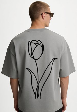 Mann trägt graues Oversize-T-Shirt mit schwarzem einzeiligem Tulpenblumen-Design auf dem Rücken, seitlich stehend, mit Sonnenbrille.