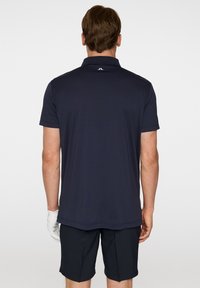 Marinblå polo t-shirt med krage och korta ärmar, med en logotyp på baksidan av nacken. Bärs med mörka shorts och en vit handske.