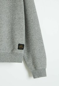 Szary sweatshirt z fakturowanej tkaniny, prążkowanymi mankietami i czarną metką logo na dolnym rękawie. Prosty design bez dodatkowych wzorów.