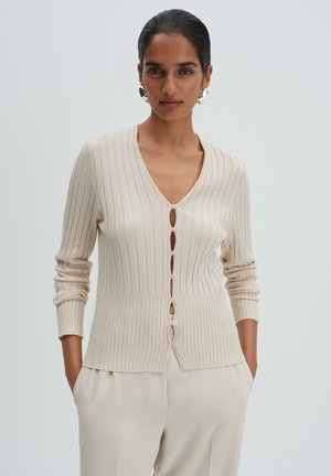 Frau, die eine cremefarbene, gerippte, geknöpfte Strickjacke mit langen Ärmeln und passende helle Hose trägt, steht mit den Händen in den Taschen.