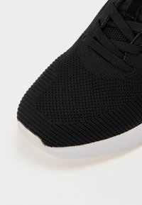 Fekete sportcipő, jellegzetes kötött felsőrésszel, bordázott mintázattal és kontrasztos fehér talppal a stabilitás és a támogatás érdekében.