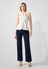 Hvit peplum topp med knyttet halslinje, i kombinasjon med mørkeblå vide jeans. Modellen har på seg gullfargede sandaler med remmer. Enkel bakgrunn.
