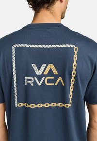 Camiseta de algodón azul marino con un gráfico que presenta el texto "VA" y "RVCA", rodeado por un diseño de cadena y cuerda en blanco y oro.