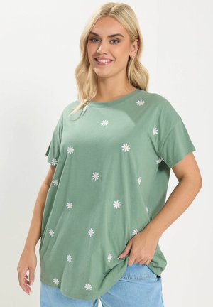 Frau trägt ein hellgrünes T-Shirt mit weißen Gänseblümchen-Drucken und hellblauen Jeans, lächelt vor einfarbigem weißem Hintergrund.