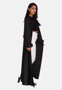 Lang sort satin frakke med brede ærmer, lagd over en kort sort top og hvide højtaljede jeans. Har åben front og glat tekstur.