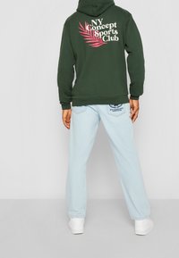 Sudadera verde con gráfico blanco y rosa "NY Concept Sports Club" en la parte posterior, combinada con vaqueros azul claros y zapatillas blancas.