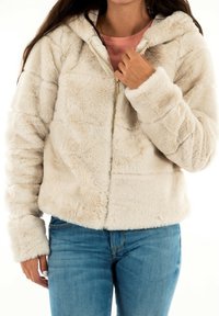 Chaqueta de piel sintética con cremallera en crema y capucha, con una textura suave y puños acanalados, combinada con una camisa rosa clara y jeans azules.