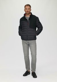 Zwarte pufferjack met een gestructureerd zwart paneel op de borst, grijze jeans en zwarte sneakers, op een model tegen een effen achtergrond.
