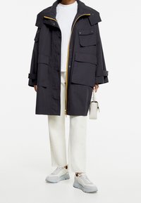 Manteau long gris foncé avec une fermeture éclair à l'avant, plusieurs grandes poches, et un accent de fermeture éclair jaune, porté sur un haut blanc et un pantalon clair.