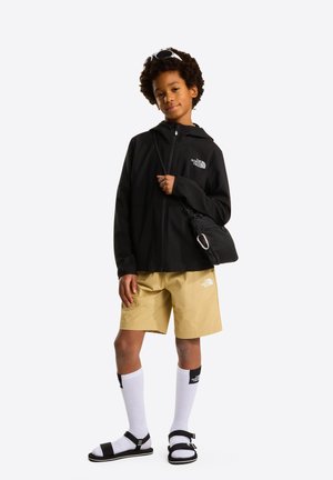 Kind steht und trägt schwarze Jacke, beige Shorts, weiße hohe Socken, schwarze Sandalen, kleine schwarze Umhängetasche und Sonnenbrille auf dem Kopf.