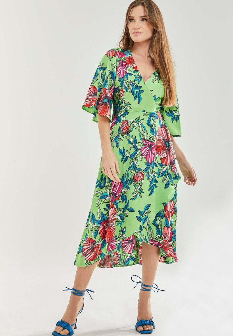 Floral wrap dress in green fabric, featuring pink and blue flowers, short flared sleeves, and a knee-length hem. Paired with blue strappy heels.
