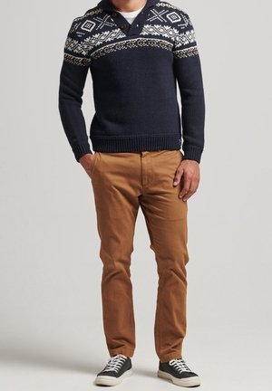Uomo che indossa un maglione blu navy a motivo, pantaloni marrone chiaro e sneakers basse nere con lacci bianchi, in piedi davanti a uno sfondo semplice.