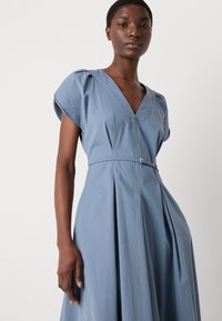 Blauwe katoenen jurk met een V-hals, geplooide rok, korte mouwen en een smal riempje met zilverkleurige details in de taille.