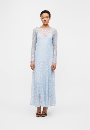 Frau steht, trägt ein langes hellblaues Spitzenkleid mit durchsichtigen langen Ärmeln und schwarzen, hochhackigen Sandalen mit Riemchen.