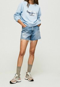 Donna che indossa un felpa leggera blu di Pepe Jeans London, pantaloni shorts in denim strappati, calze beige e sneakers multicolore, in piedi con le mani in tasca.